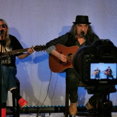 Das Duo sitzt auf Barhockern, sie spielen beide Gitarre und singen. Im Vordergrund ist eine Kamera, die gerade aufnimmt. 