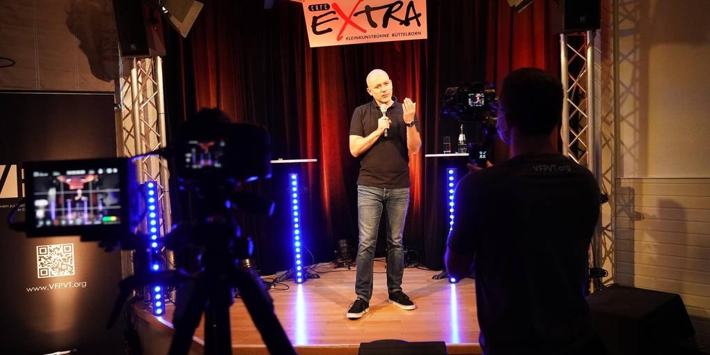 Frank Fischer stehend auf der Bühne. Ein Kameramann filmt ihn.