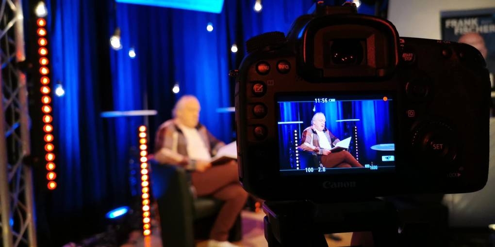 Jürgen Leber in einem Sessel auf der Bühne. Im Vordergrund ist eine Kamera mit Display.