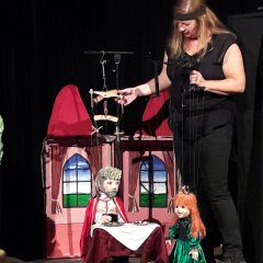 Eine Frau rechts im Bild mit zwei Marionetten. Die Puppen stellen einen König und eine Prinzessin vor einem Schloss dar.