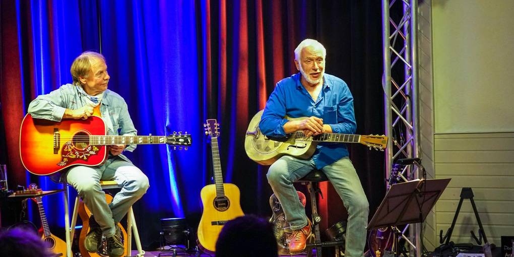 Rainer sitzt links und Bodo rechts. Beide halten eine Gitarre in der Hand. Bodo erklärt etwas.
