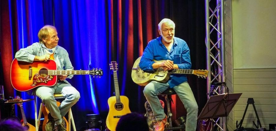 Rainer sitzt links und Bodo rechts. Beide halten eine Gitarre in der Hand. Bodo erklärt etwas.
