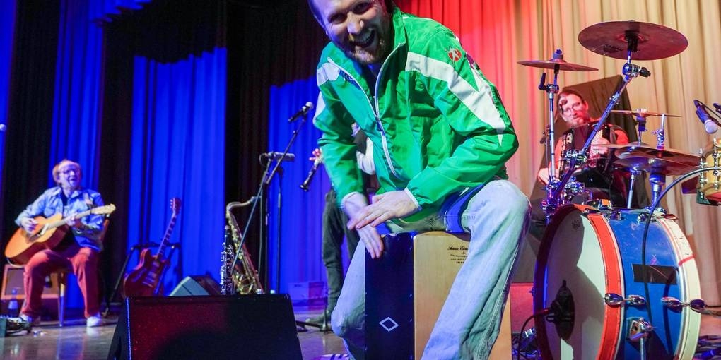 Der Frontsänger lacht in die Kamera. Es sind verschiedene Instrumente zu sehen. 