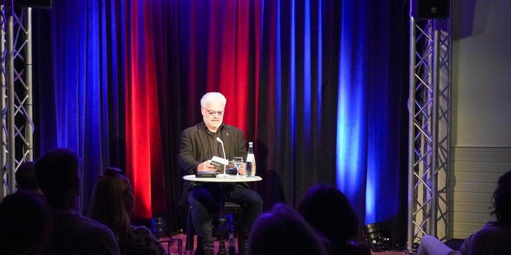 Kibler bei seiner Lesung im Café Extra. Er sitzt an einem Tisch auf der Bühne und liest vor.