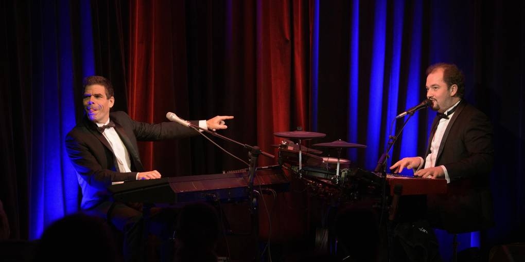 Zwei Musiker im Anzug sitzen sich gegenüber auf der Bühne. Beide spielen Klavier und singen dazu.