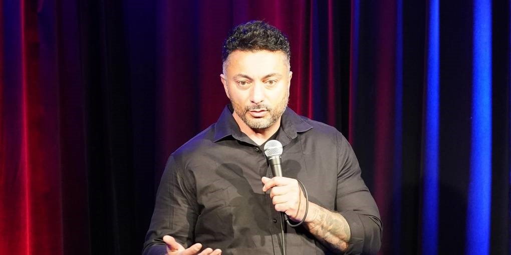 Cebe steht auf der Bühne und spricht zum Publikum, dabei hält er ein Mikrophon in der Hand.